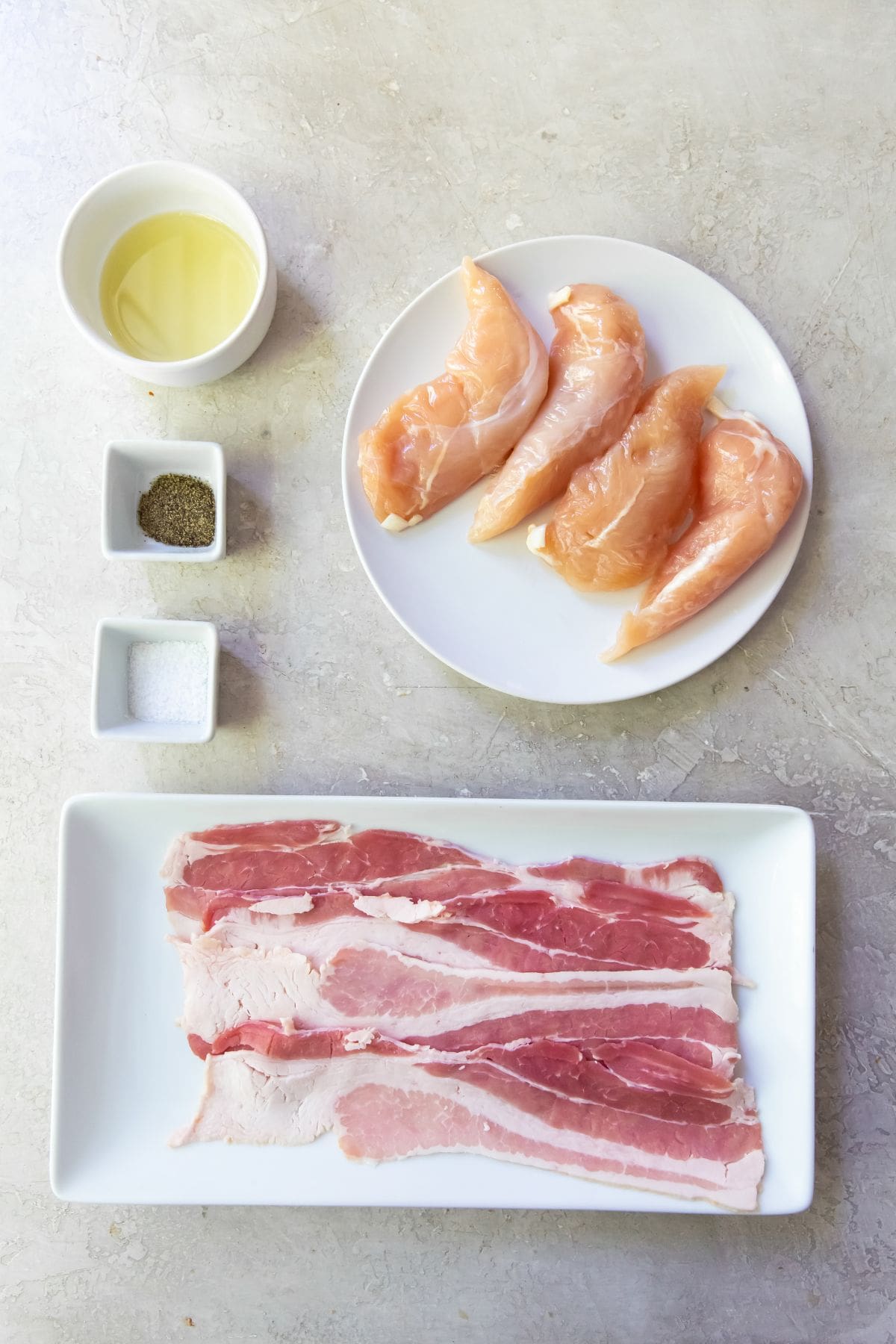 Gray background with ingredients lined up. bacon on a white plate. chicken tenders on a white plate. Salt and black pepper in a white side dish avocado oil in white side dish