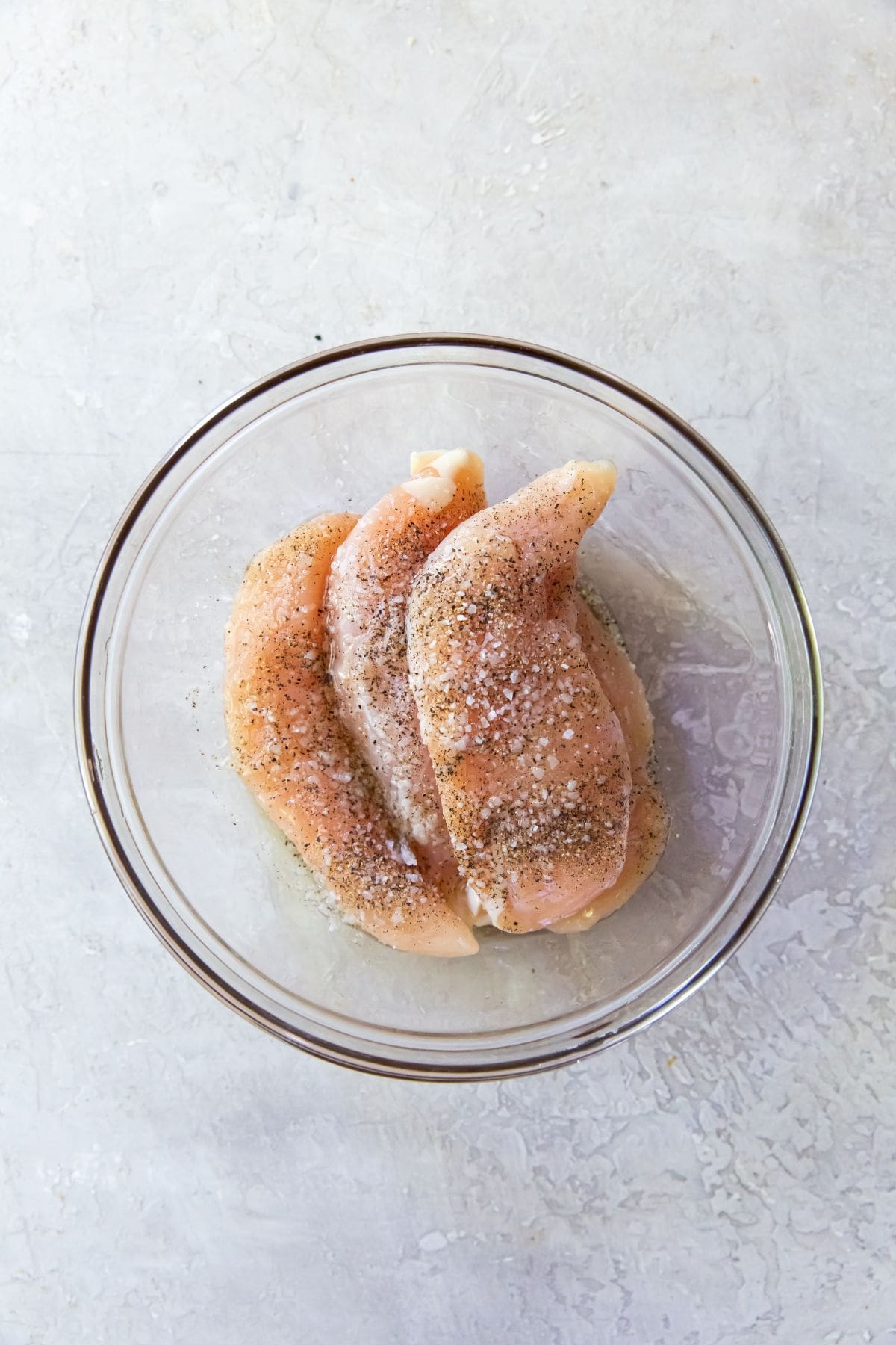 Glass mixing bowl with chicken tenders coated in salt, black pepper, and avocado oil