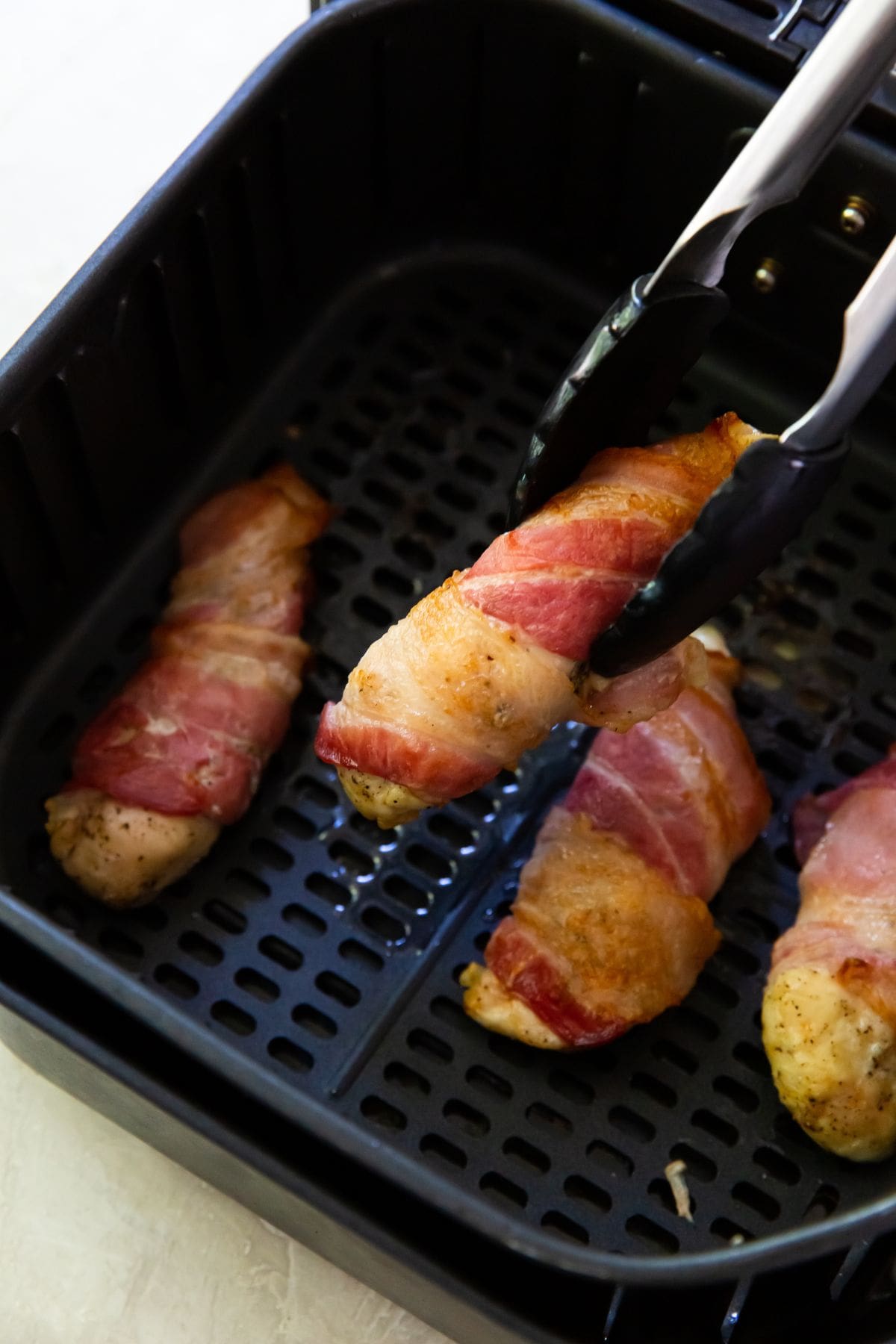 Black Air Fryer with cooked bacon wrapped chicken tenders tongs holding one tender