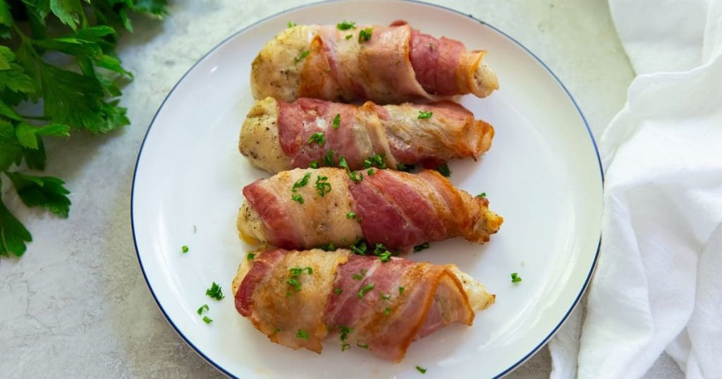 bacon wrapped chicken tenders on a white plate. parsley top left corner. white towel on side