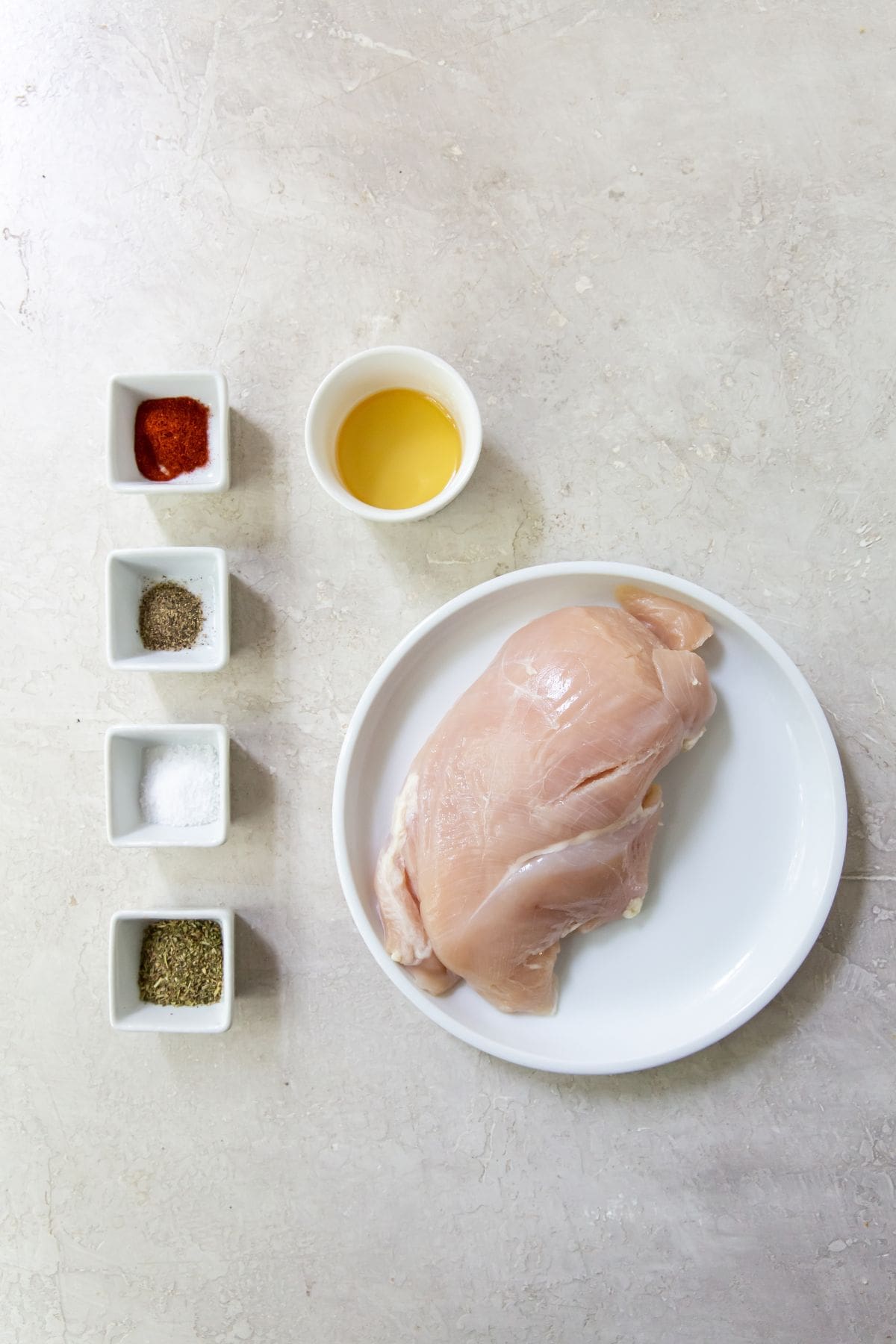 Gray background with ingredients. on the left 4 square dishes with paprika, black pepper, salt, and Italian seasonings. In the middle a white bowl of avocado oil underneath a chicken breast 