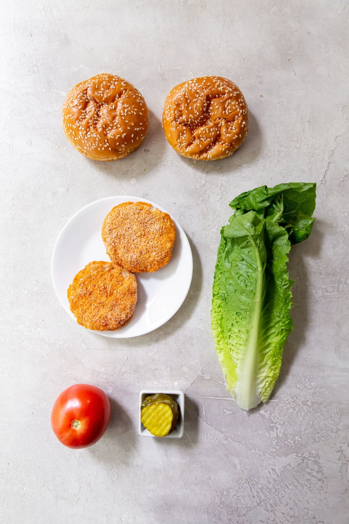 Hamburger buns at the top. Frozen chicken patties on a white plate. A piece of lettuce next to it. A whole tomato and pickles in a square serving dish