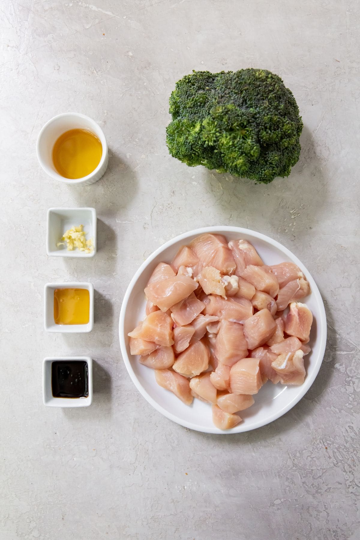 Gray background with white serving dishes broccoli in the background. Raw chicken on white plate. Soy sauce, honey, and minced garlic in square white dishes. Avocado oil in white circle dish