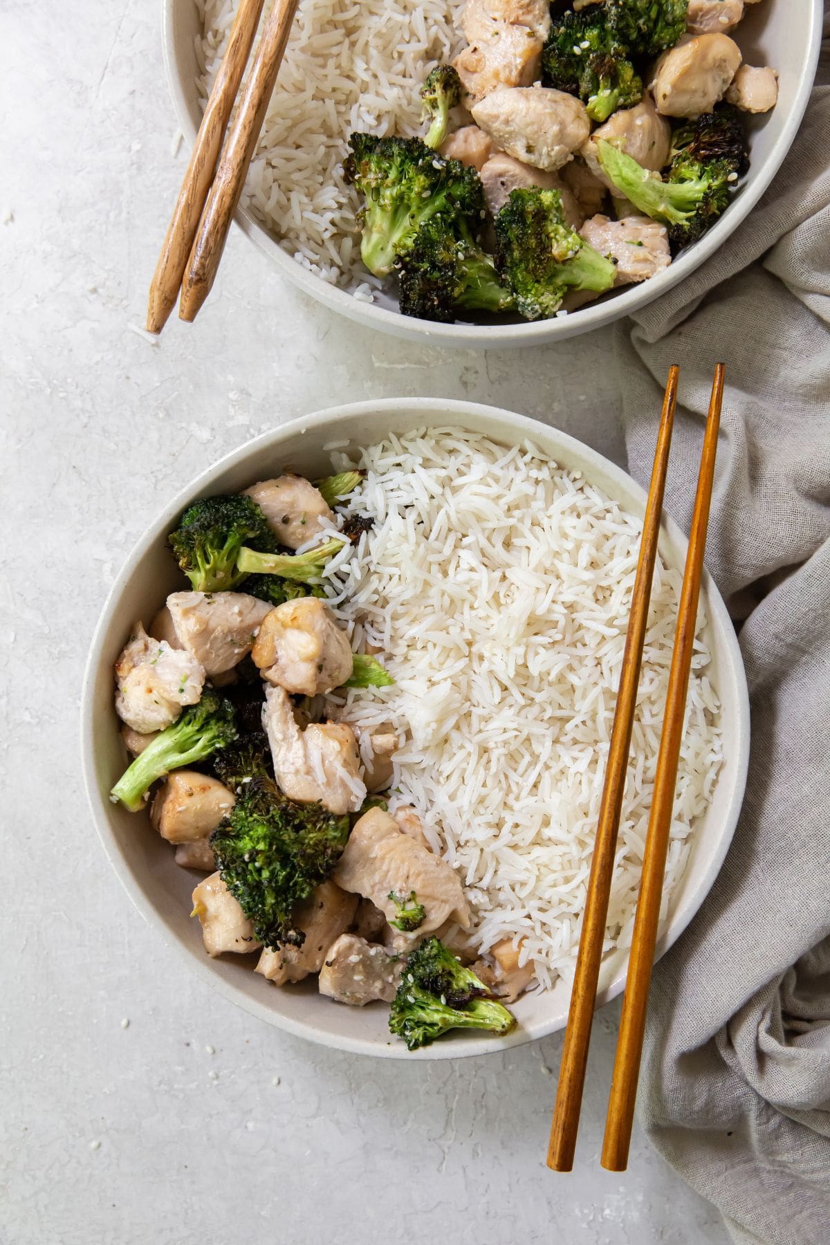 Gray background with white bowl that has chicken and broccoli. White rice on the side. Brown chopsticks on side