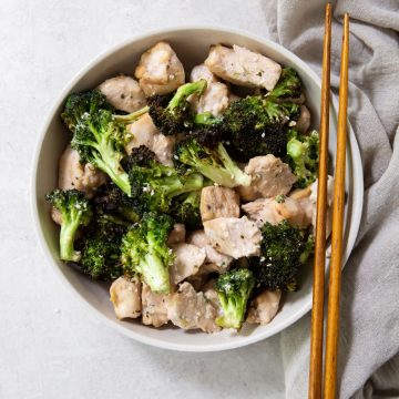 white bowl with chicken and broccoli inside it. brown chopsticks on the side gray towel on the side