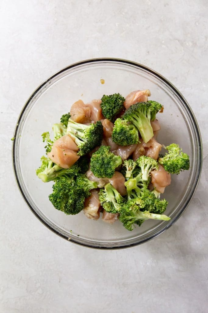 Gray background. Glass bowl has chicken and broccoli tossed in ingredients