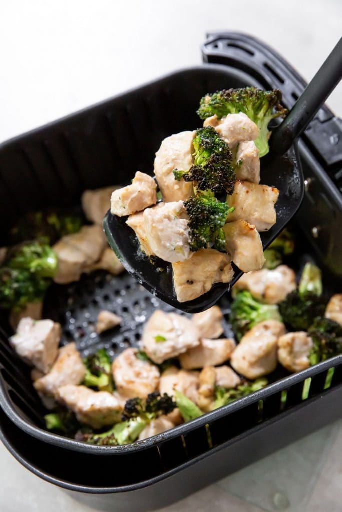 Black air fryer with finished chicken and broccoli. Holding a spatula over the air fryer