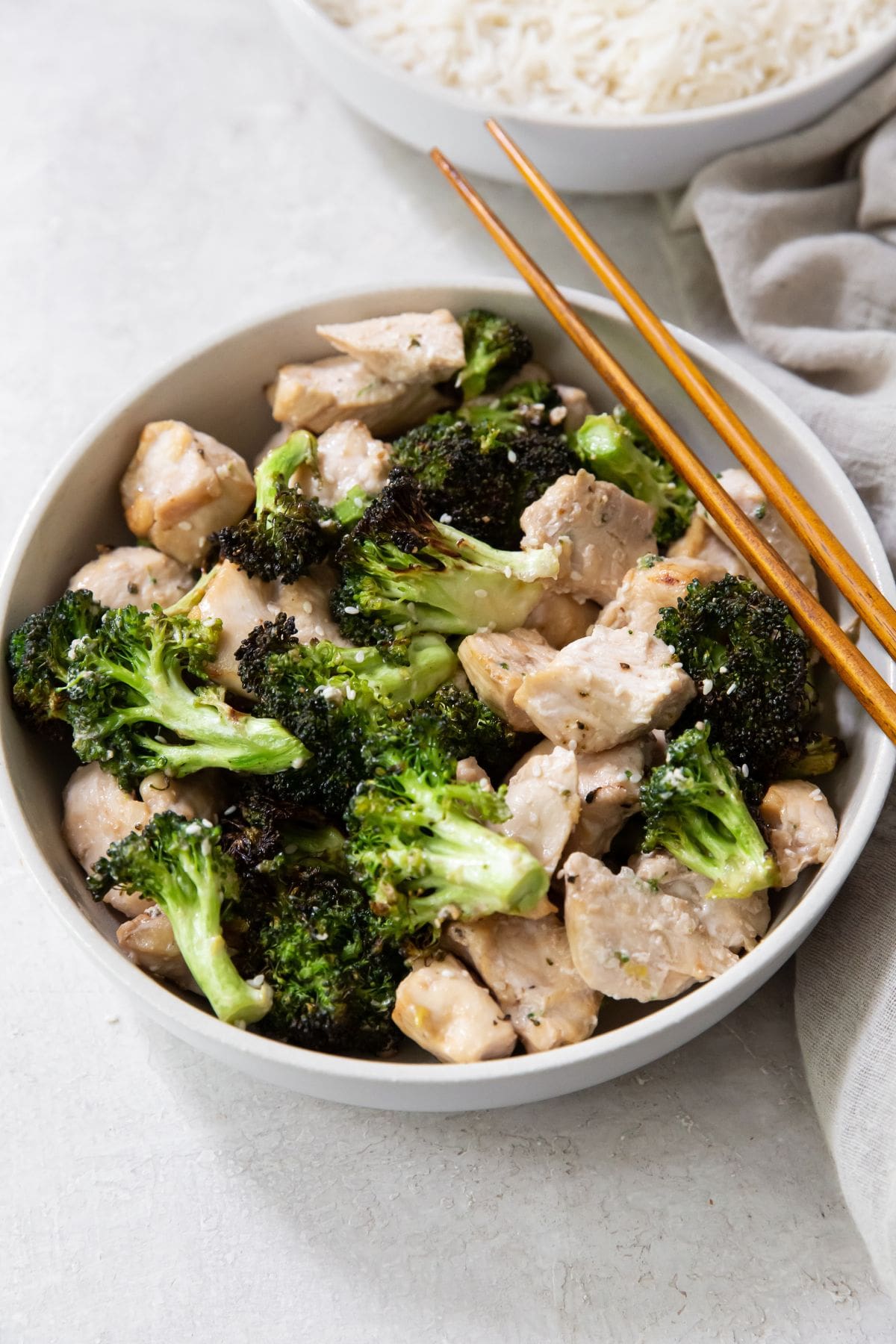 Gray background with white bowl filled with Broccoli and chicken. brown chopsticks on the right side with gray napkin on the side