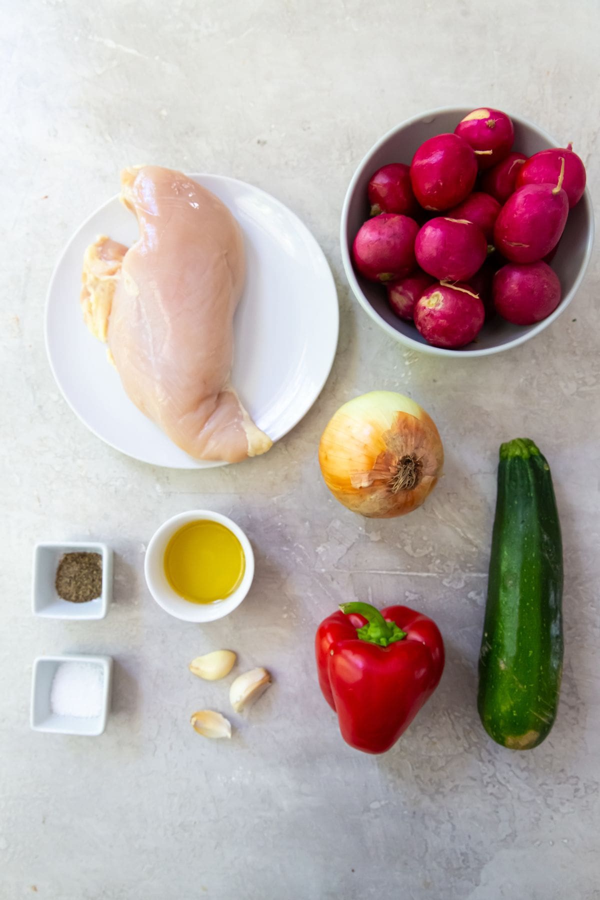 Gray background with ingredients lined up. Radishes in a bowl chicken breast on a plate onion pepper zucchini by their selves. salt pepper avocado oil in a bowl. garlic