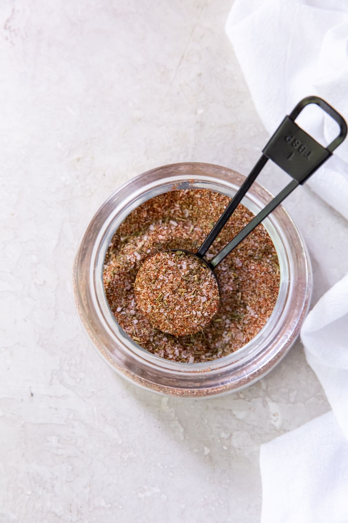 Glass jar with dry rub seasoning tablespoon inside jar. white towel next to it