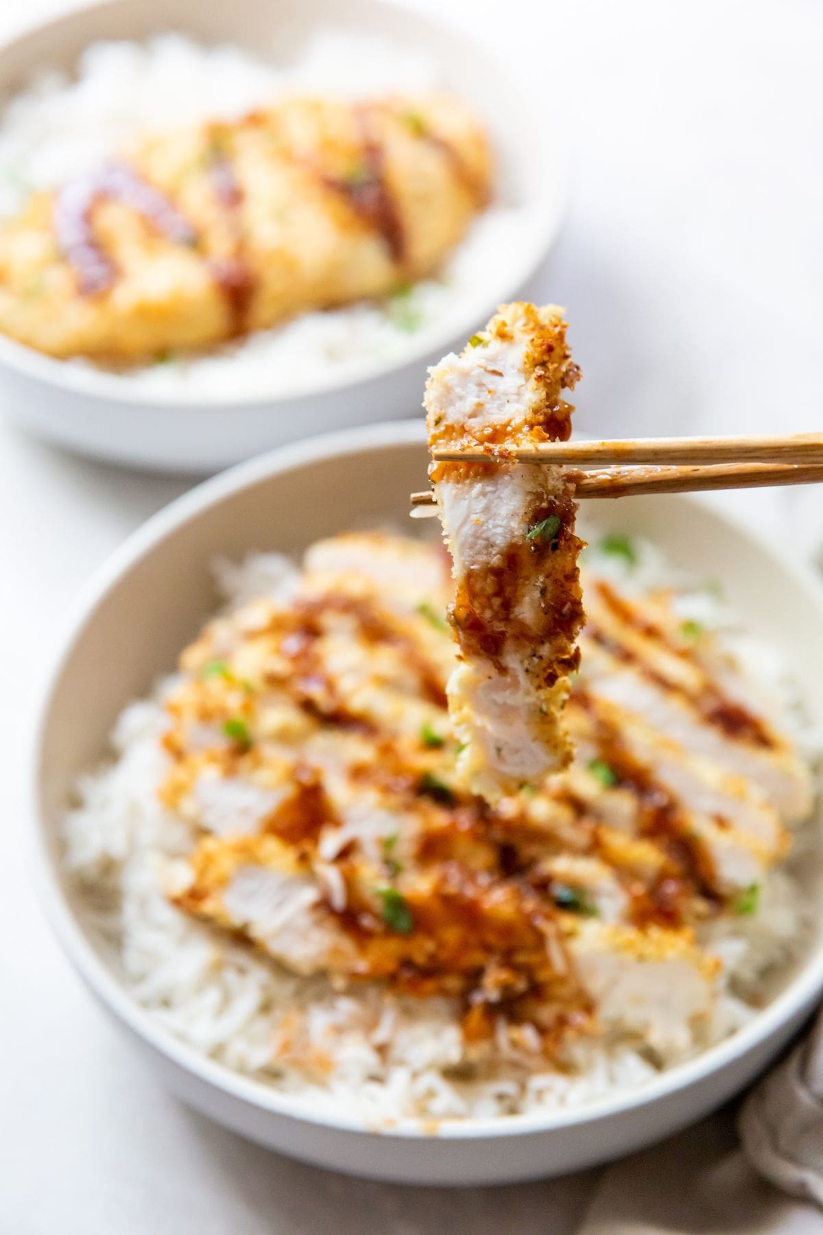 white bowl with air fryer chicken katsu under a bed of rice chopsticks holding a piece of chicken