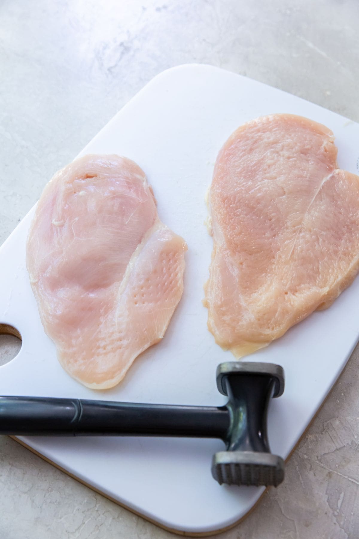 white cutting board with chicken breast meat mallet next to it