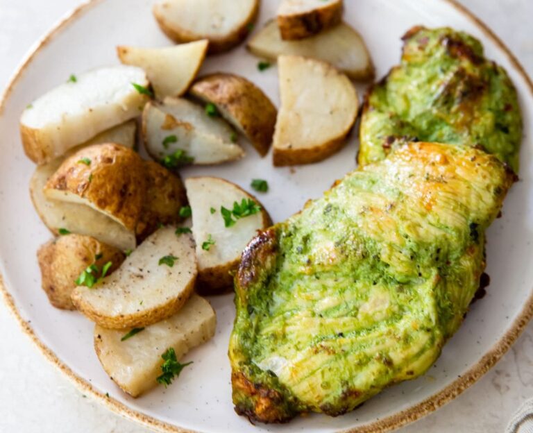 A white plate with seasoned roasted potato wedges and a piece of green herb-marinated pesto chicken, cooked to perfection in the air fryer, garnished with chopped parsley.