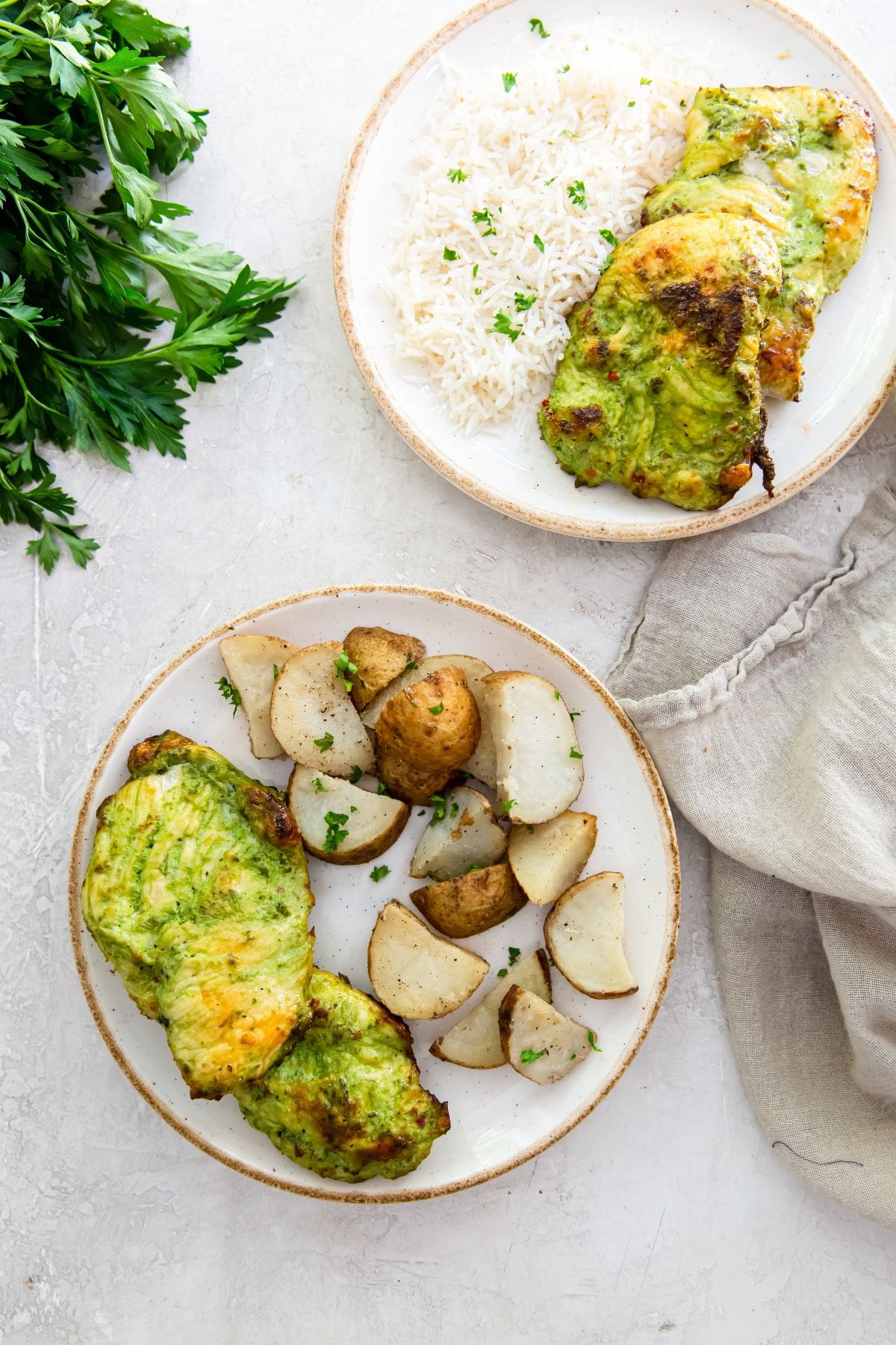 white plate with chicken and rice. another plate in the background with chicken and potatoes on it. parsley in the back. green towel next to chicken and rice