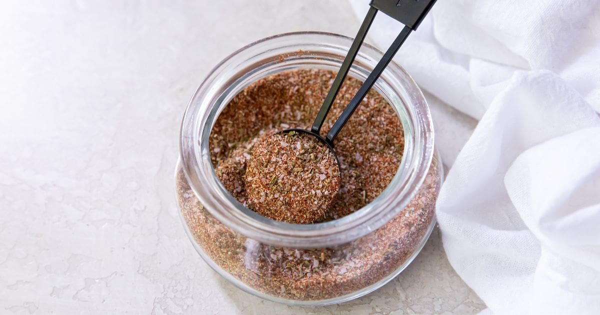 Glass jar with dry rub seasoning tablespoon inside jar. white towel next to it