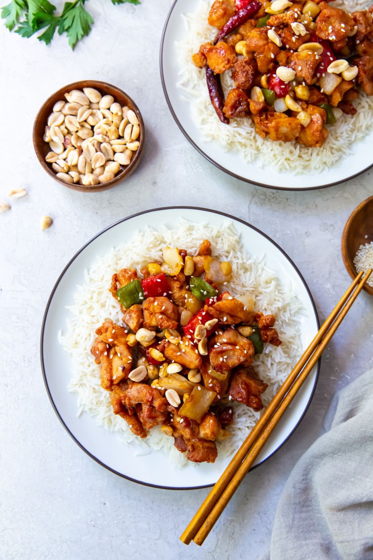 two plates with rice and kung pao chicken on it. parsley in the back. a bowl with peanuts and sesame seeds. Gray towel next to it. Chopsticks on one plate