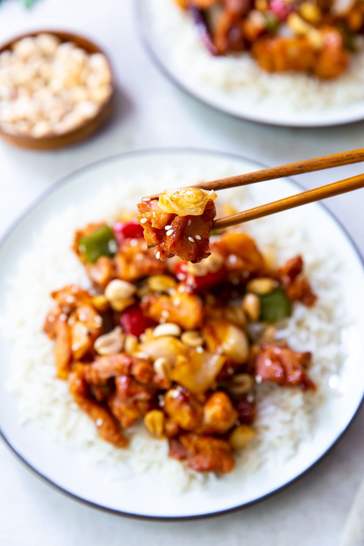 two plate with rice and kung pao chicken on it. bowl of peanuts next to it. chopsticks holding a piece of chicken and onion