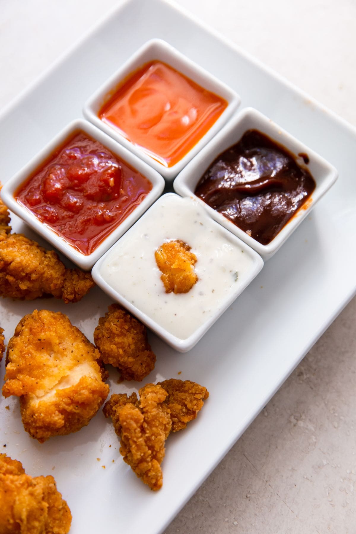 white plate with popcorn chicken on it buffalo, ketchup, bbq, ranch. one popcorn chicken dipped in ranch.