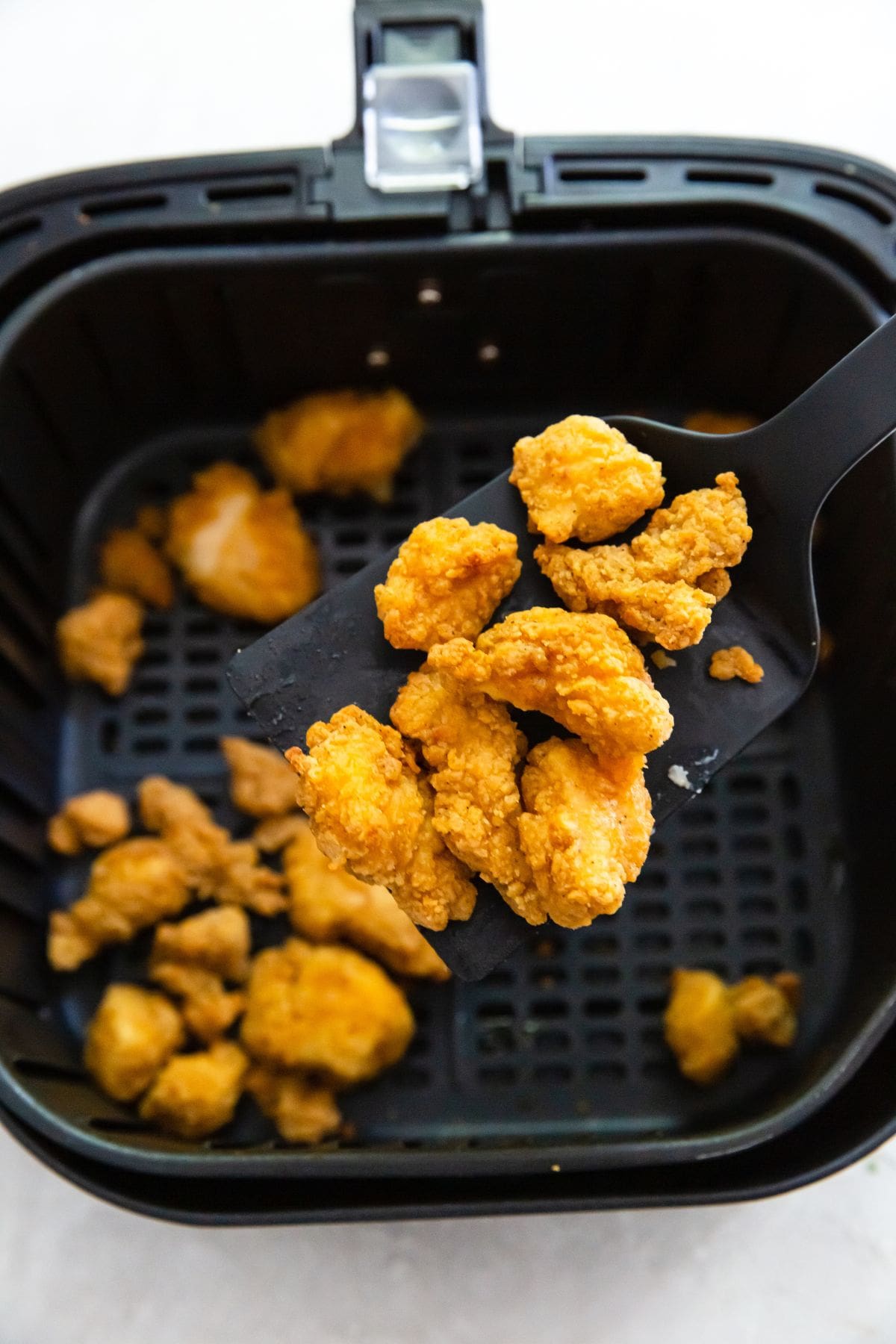 black air fryer basket with cooked popcorn chicken. Some popcorn chicken on a spatula being held