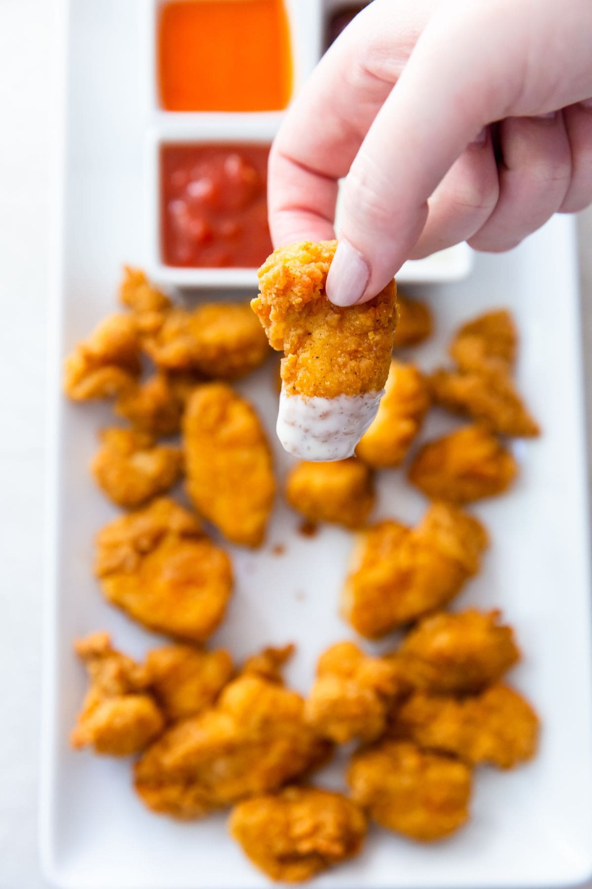 white plate with popcorn chicken on it buffalo, ketchup, bbq, and ranch in the back. One popcorn chicken with ranch held over the top.