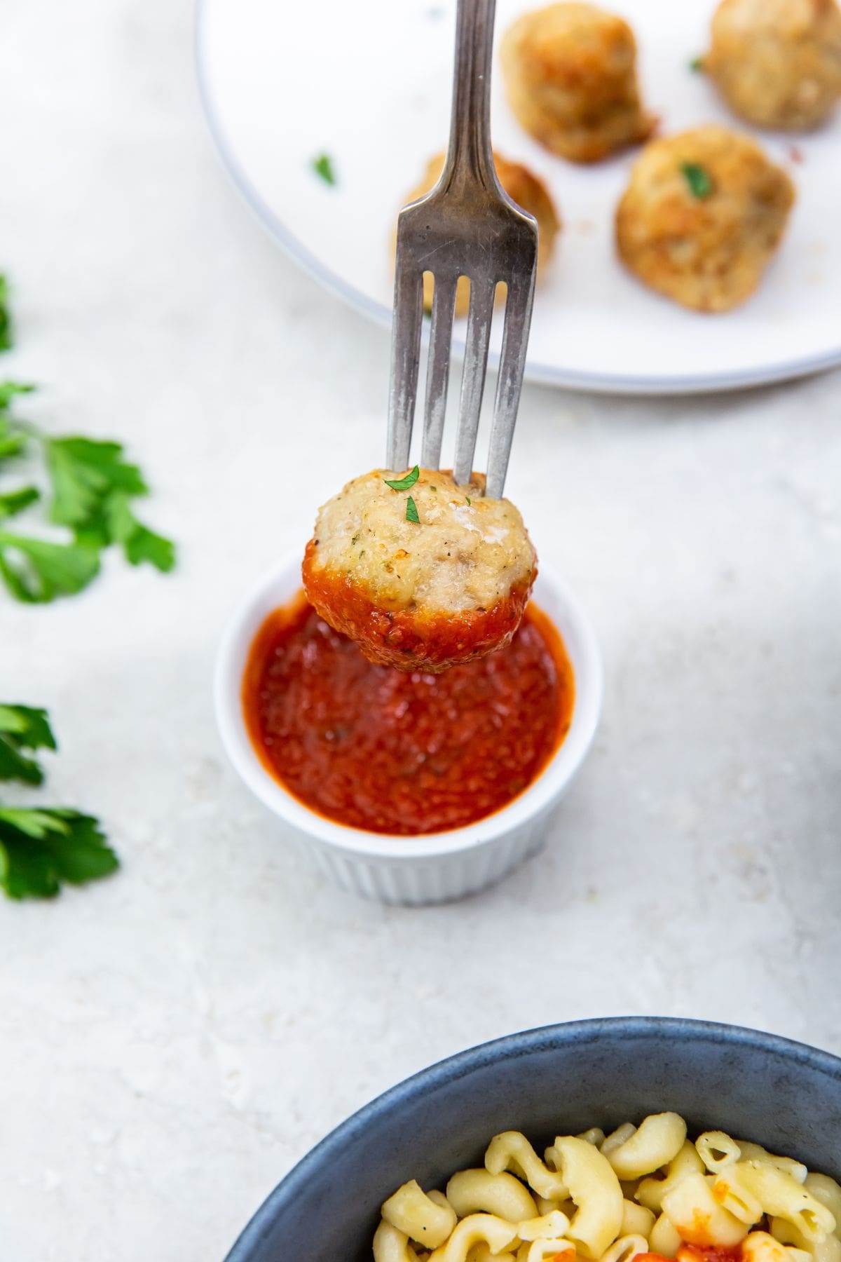 meatball dunked into the sauce with a fork. plate with meatballs inn the back.parsley on the side