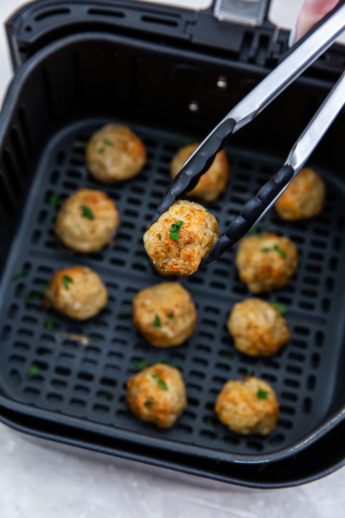 black air fryer with cooked ground chicken with parsley on top. tongs holding up one meatball