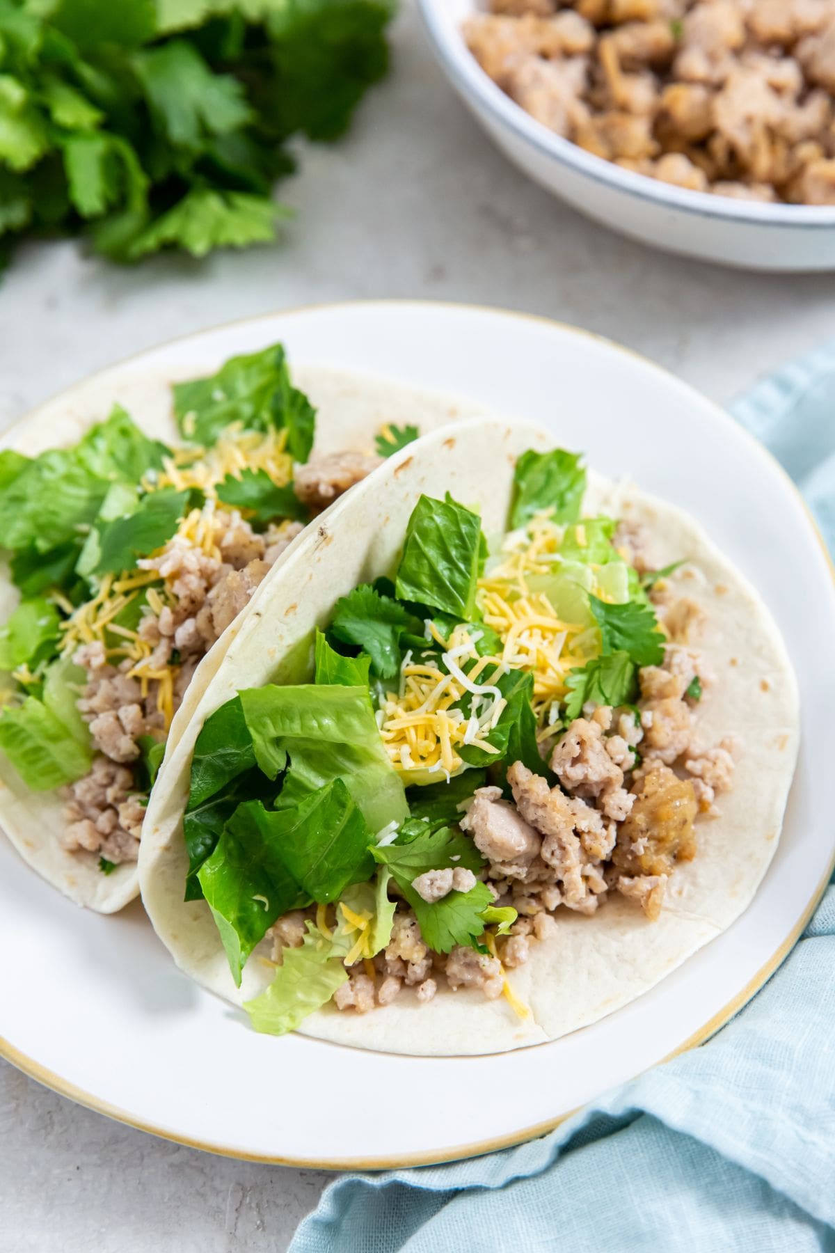 two chicken tacos on a plate. bowl of chicken in the back. parsley and blue towel next to them