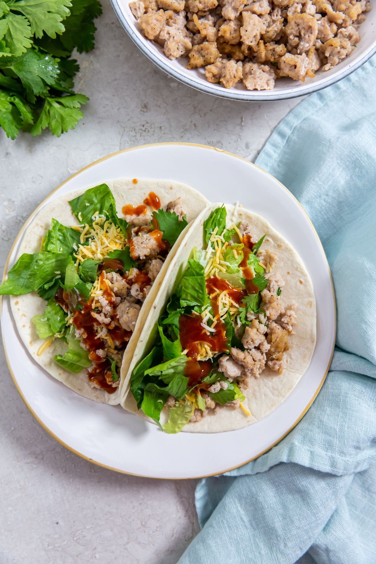 two chicken tacos on a plate. bowl of chicken in the back. parsley and blue towel next to them