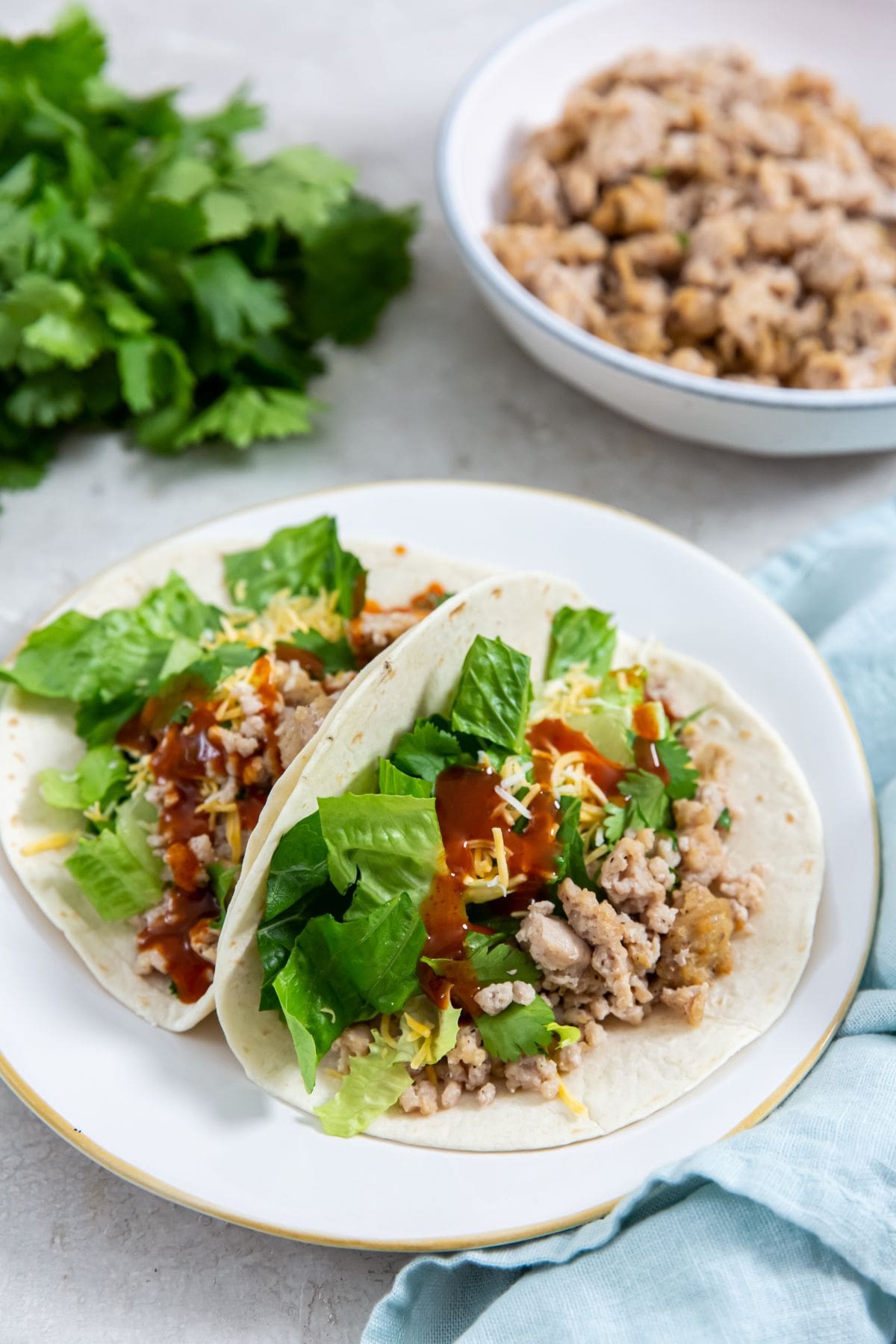 two chicken tacos on a plate. bowl of chicken in the back. parsley and blue towel next to them