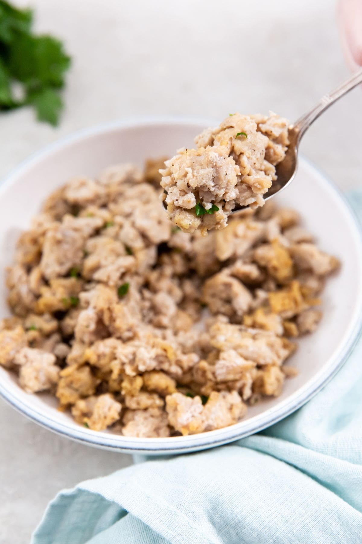 white bowl with chicken inside it. spoon being held over the top of it. blue towel and parsley in the back