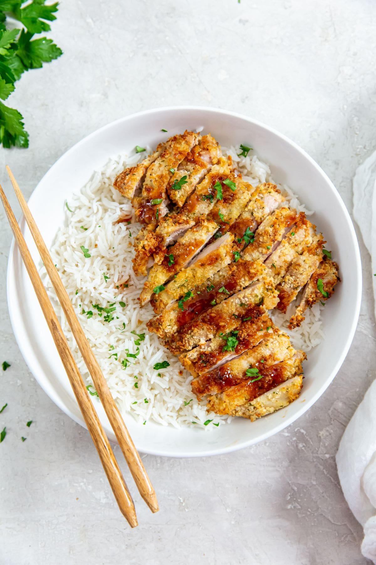 chicken and rice in a white bowl. chopsticks on the side of the bowl parsley and white towel in the back