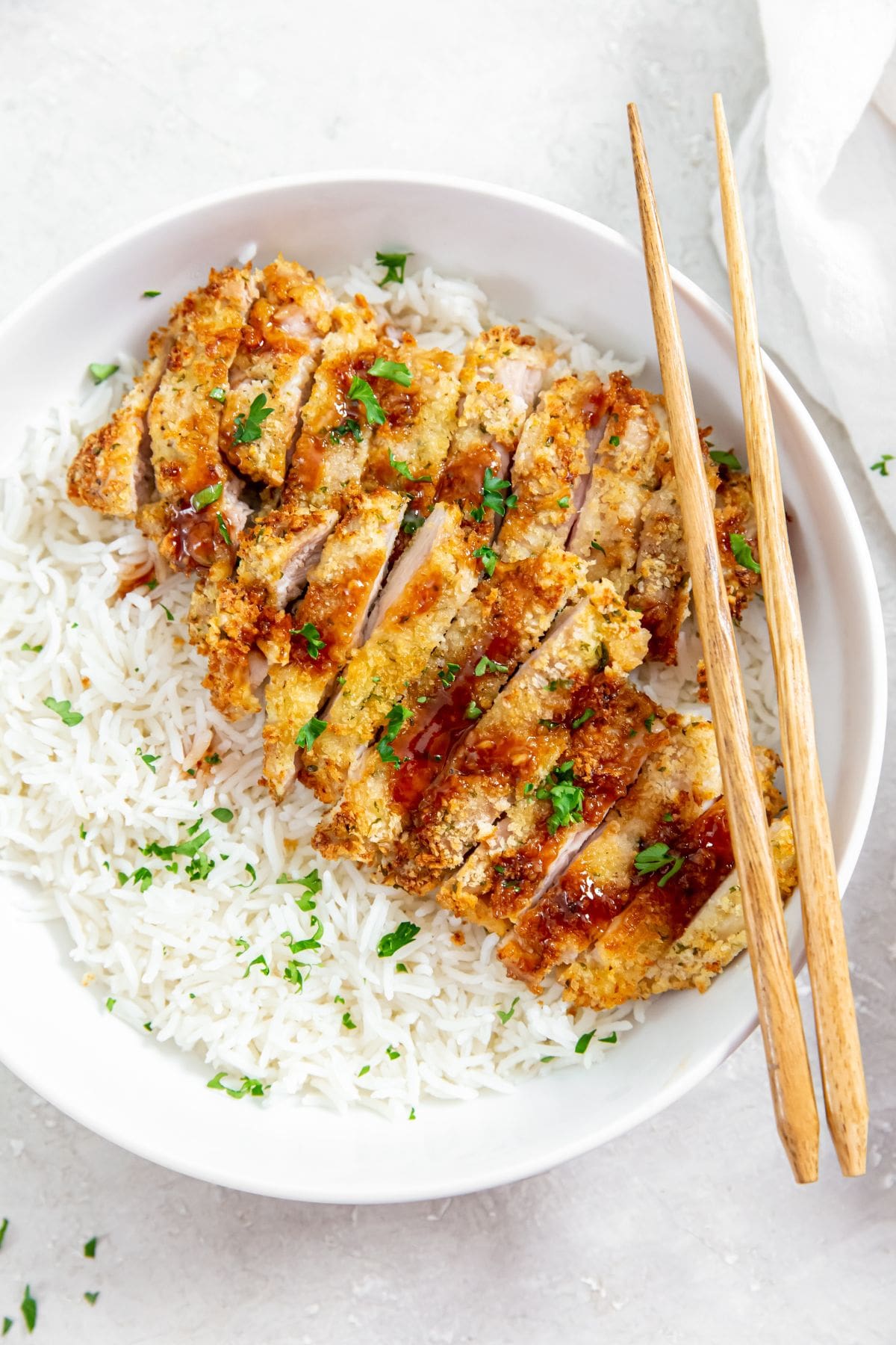 chicken and rice in a white bowl. chopsticks on the side of the bowl parsley and white towel in the back