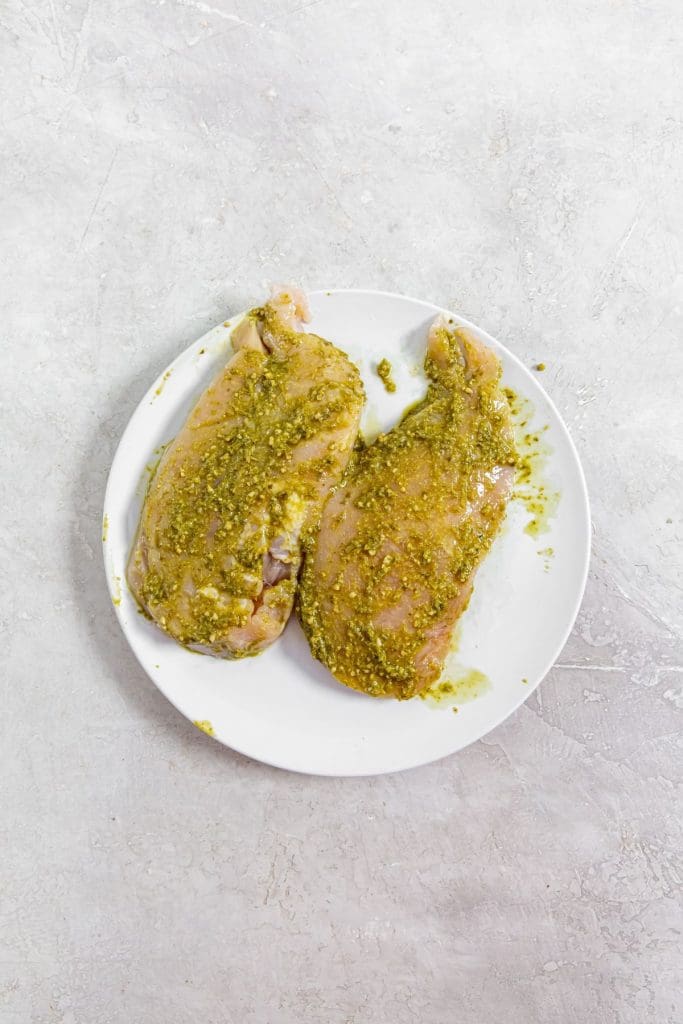 white plate with raw chicken and pesto spreader all over the chicken breast