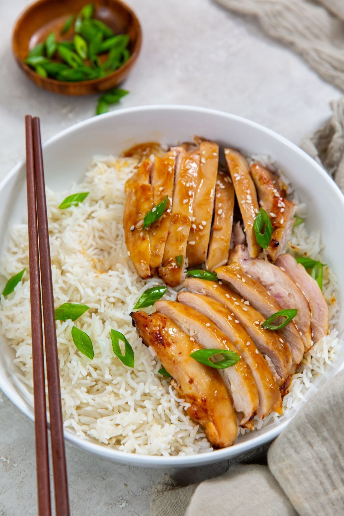 white bowls with rice and chicken thighs inside. Green onions on top. Gray towel in the back. sesame seeds and green onion in a brown bowl chopsticks on bowl