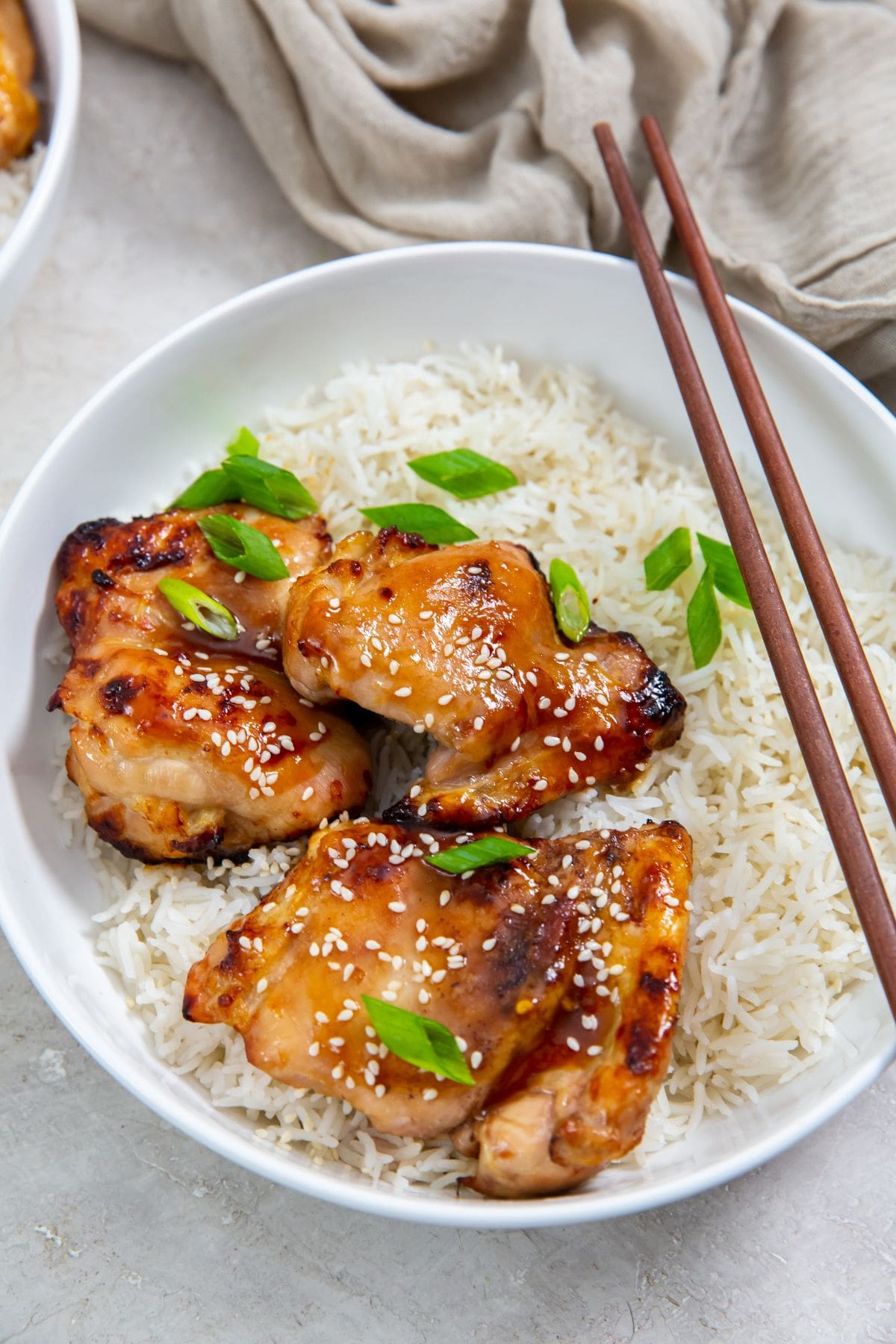 white bowls with rice and chicken thighs inside. Green onions on top. Gray towel in the back. sesame seeds and green onion in a brown bowl chopsticks on bowl