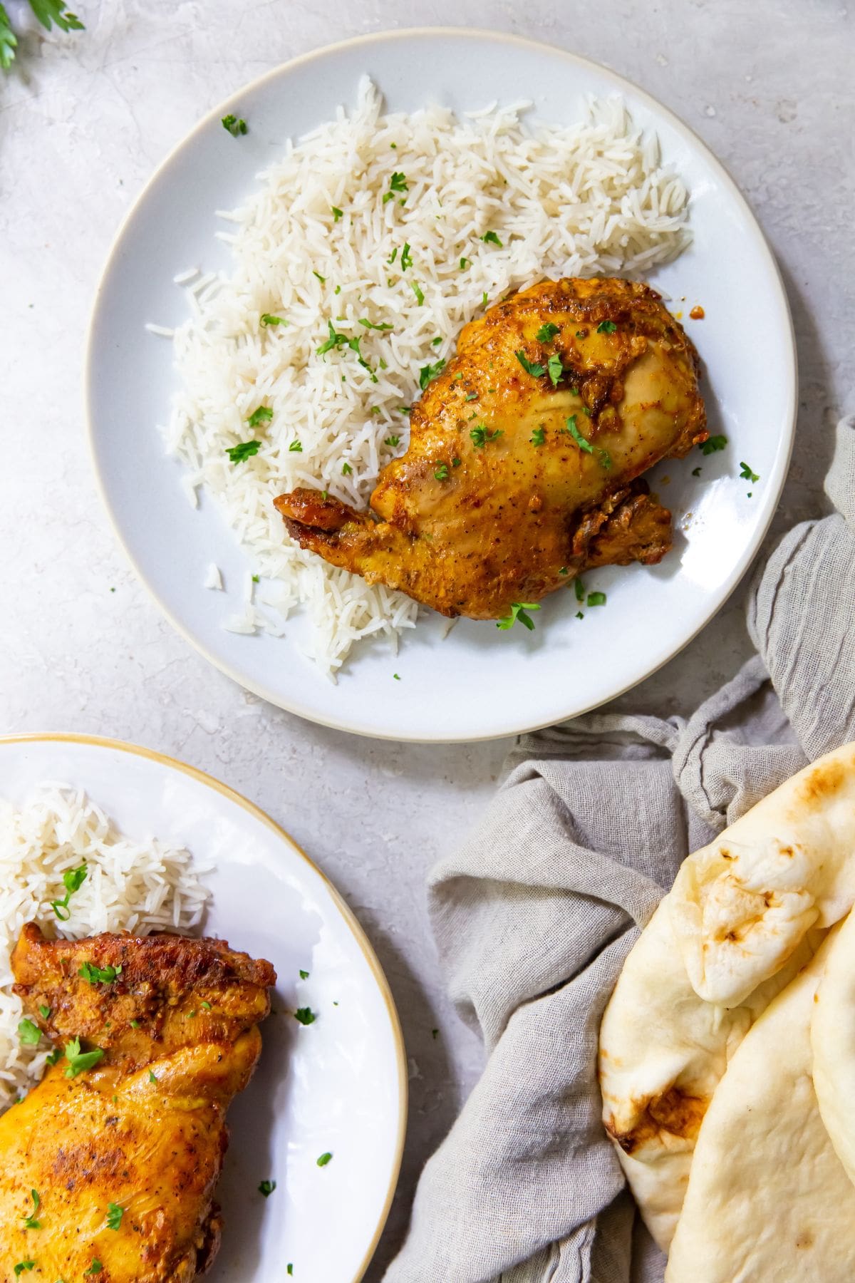 white plate with chicken thighs and rice on it parsley sprinkled on top. Naan on the side gray towel and parsley in the back