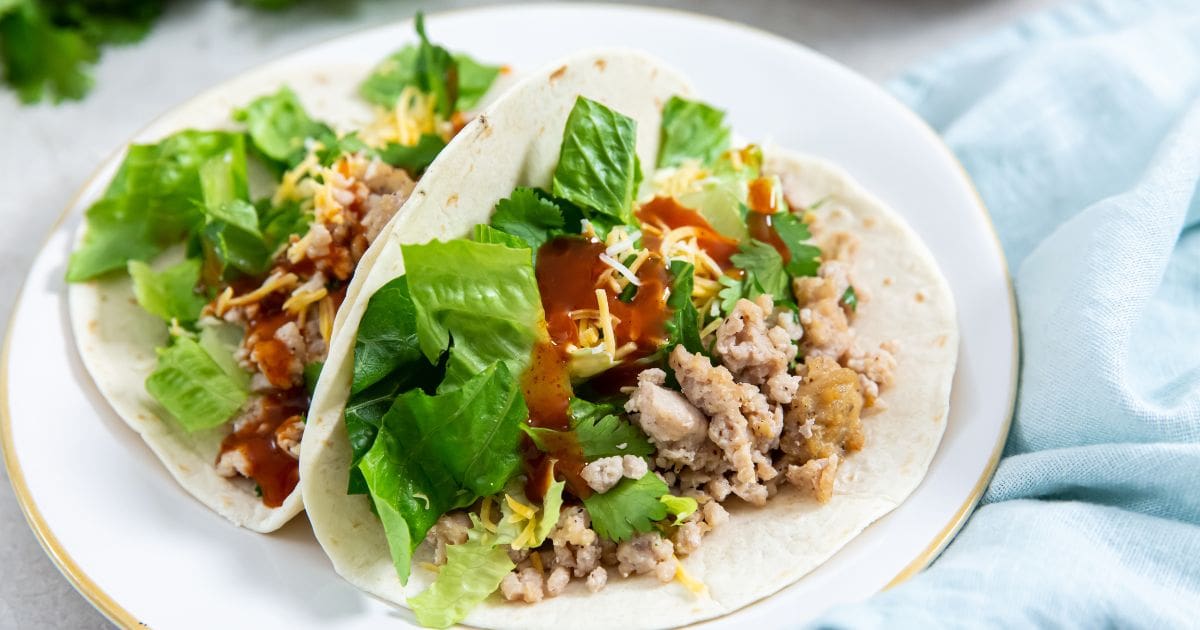 two chicken tacos on a plate. bowl of chicken in the back. parsley and blue towel next to them