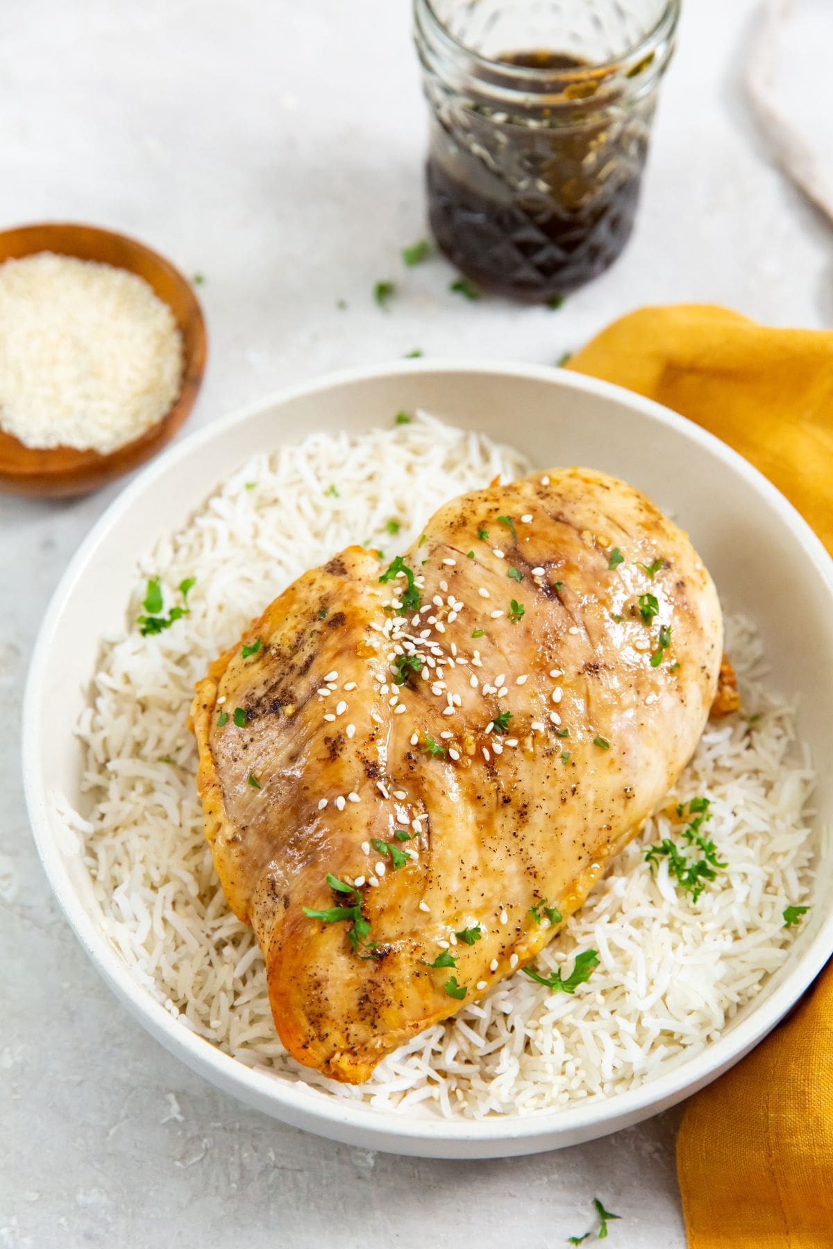 chicken on top of a bed of rice. parsley sprinkled on top. orange towel next to it. sesame seeds and glaze in thee back