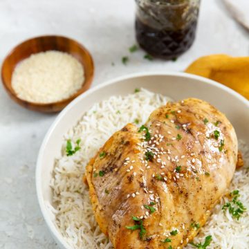 chicken on top of a bed of rice. parsley sprinkled on top. orange towel next to it. sesame seeds and glaze in thee back