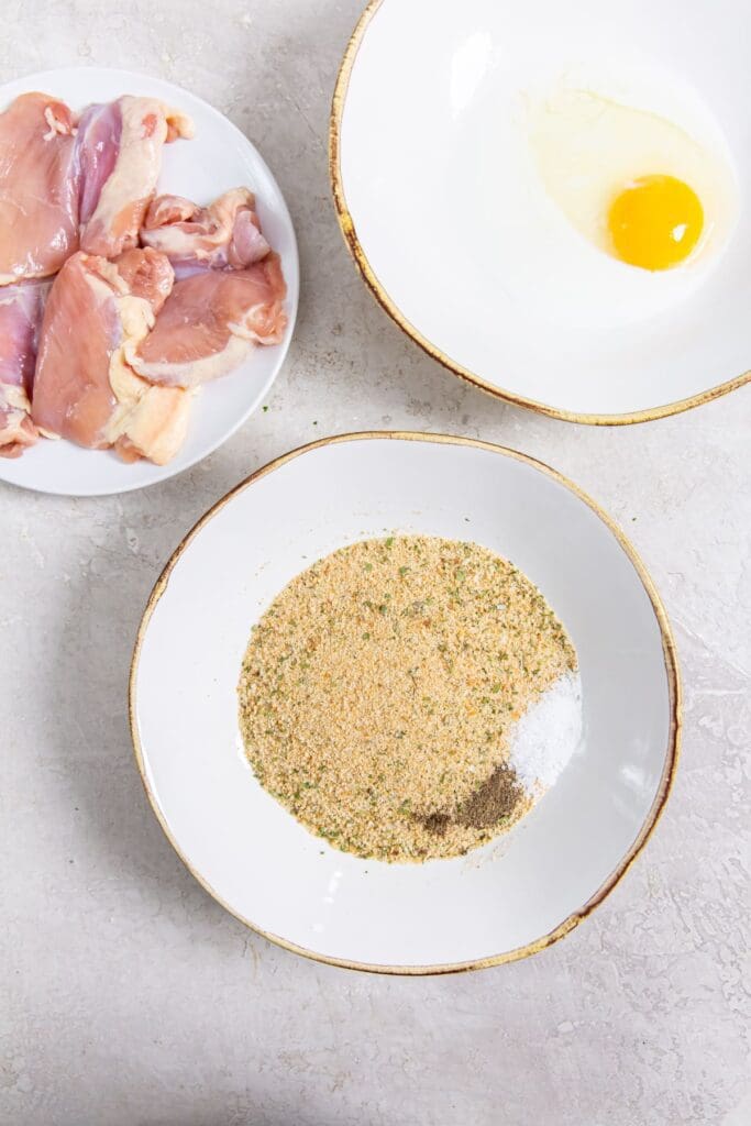 three bowls one with chicken thighs one has dry ingredients the other has egg and milk