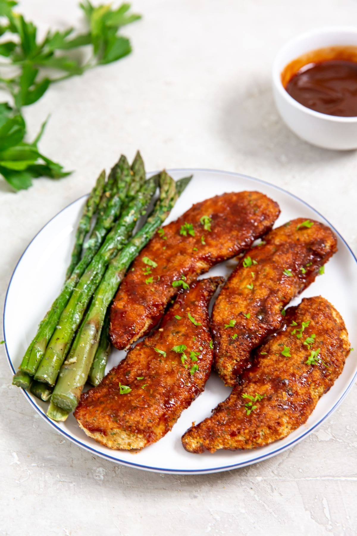 white plate with chicken and asparagus on it parsley and bbq sauce in the back