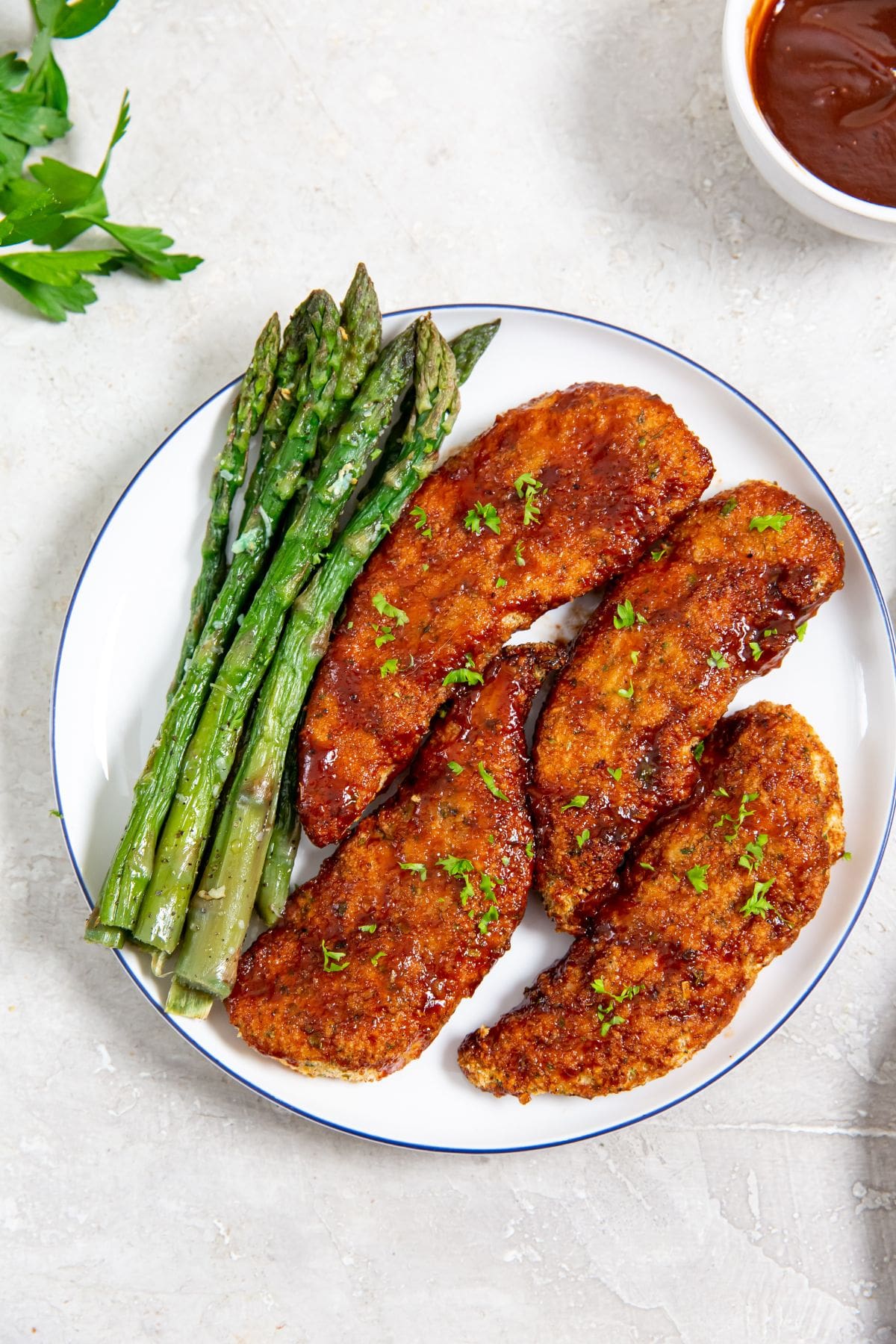 white plate with chicken and asparagus on it parsley and bbq sauce in the back