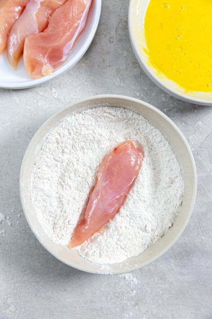 three bowls one with chicken, one with flour and another with egg. chicken tender in flour