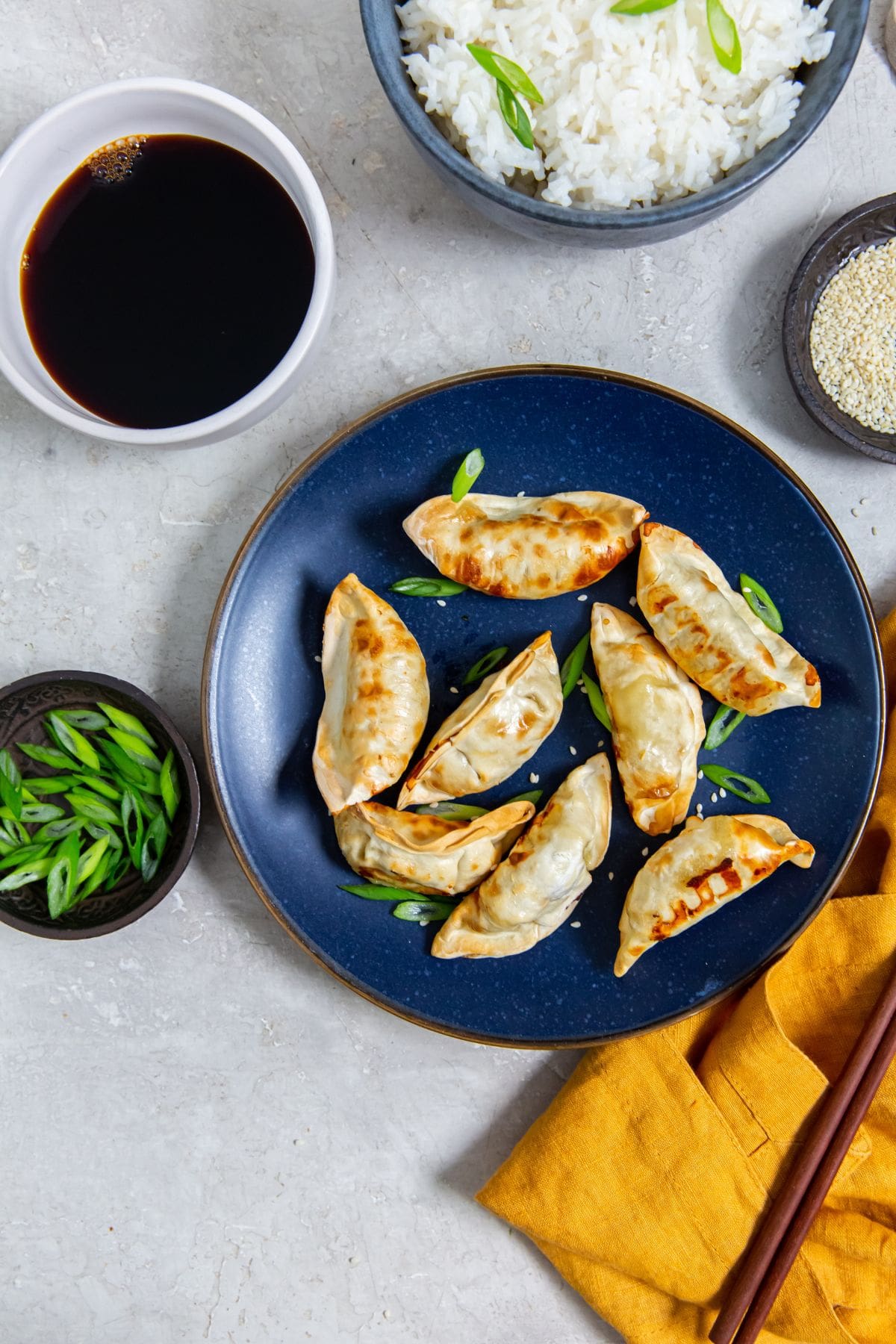 Blue plate with potstickers and scallions on it. chopsticks on the side of the plate. orange towel next to it.rice in the back