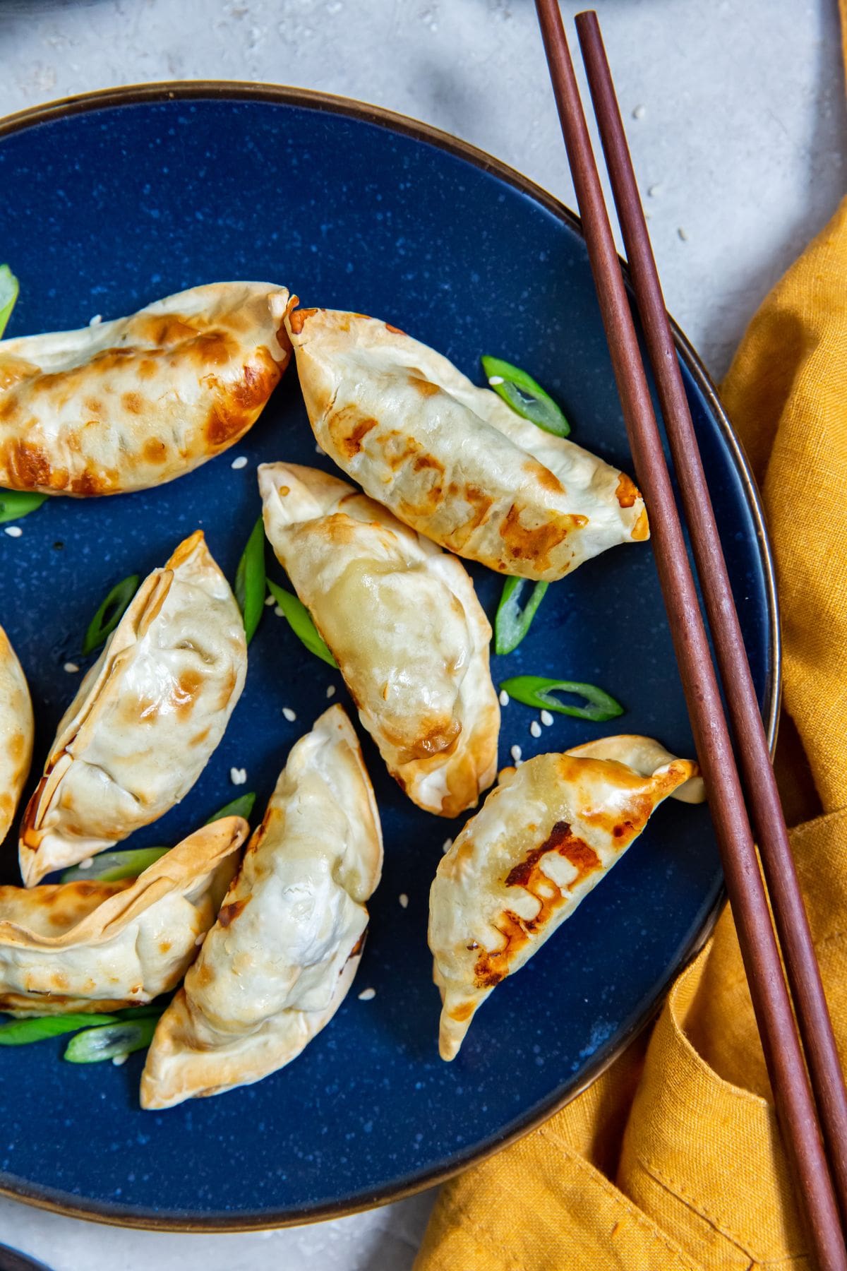 Blue plate with potstickers and scallions on it. chopsticks on the side of the plate. orange towel next to it.