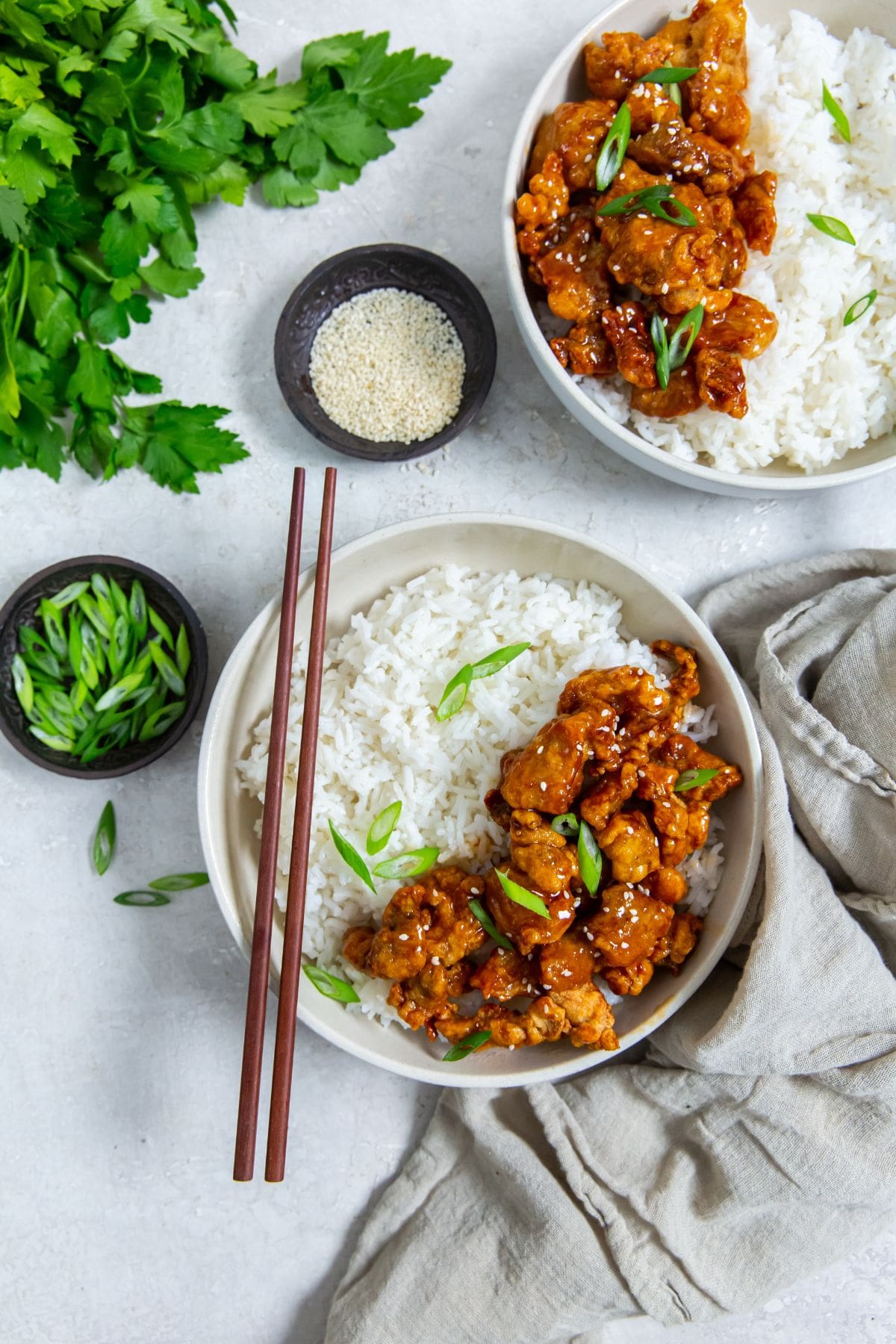 white bowl with chicken and rice inside. scallions on top. Gray towel next to it. scallions and sesame seeds in the back. chopsticks on the bowl