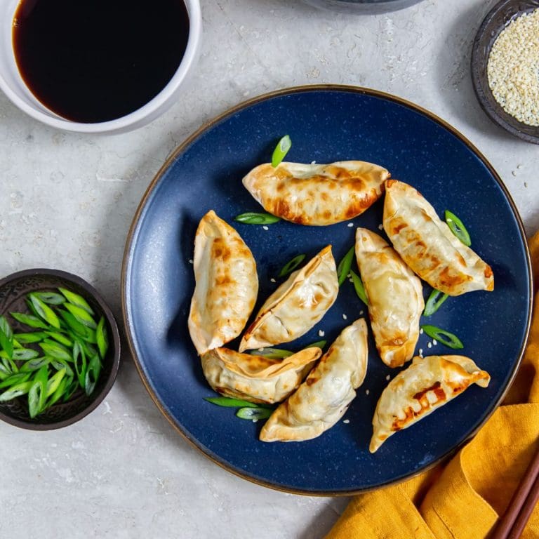 Blue plate with potstickers and scallions on it. chopsticks on the side of the plate. orange towel next to it.rice in the back