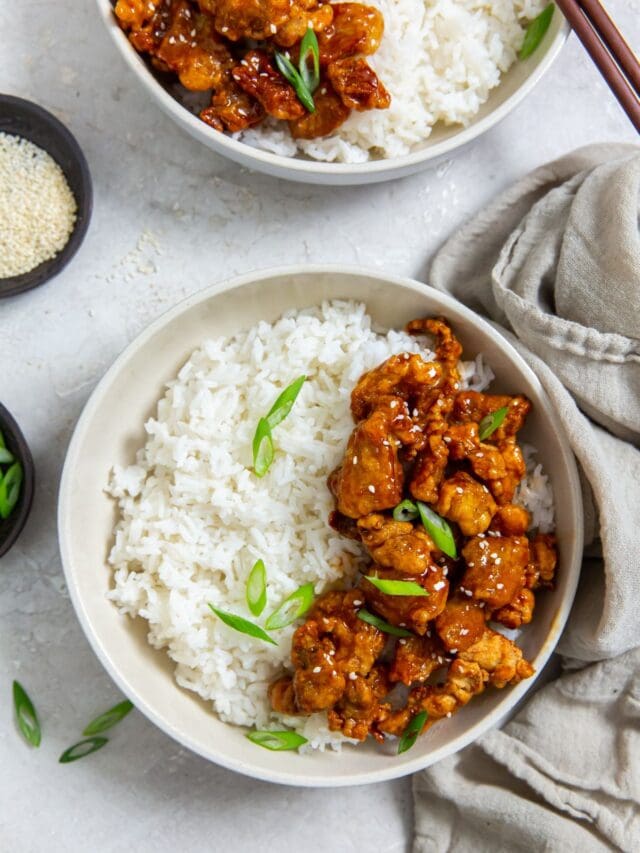 white bowl with chicken and rice inside. scallions on top. Gray towel next to it. scallions and sesame seeds in the back. chopsticks on the bowl