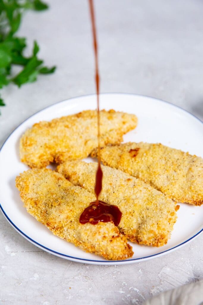 white plate with chicken on it. katsu sauce being poured on top. parsley I the back