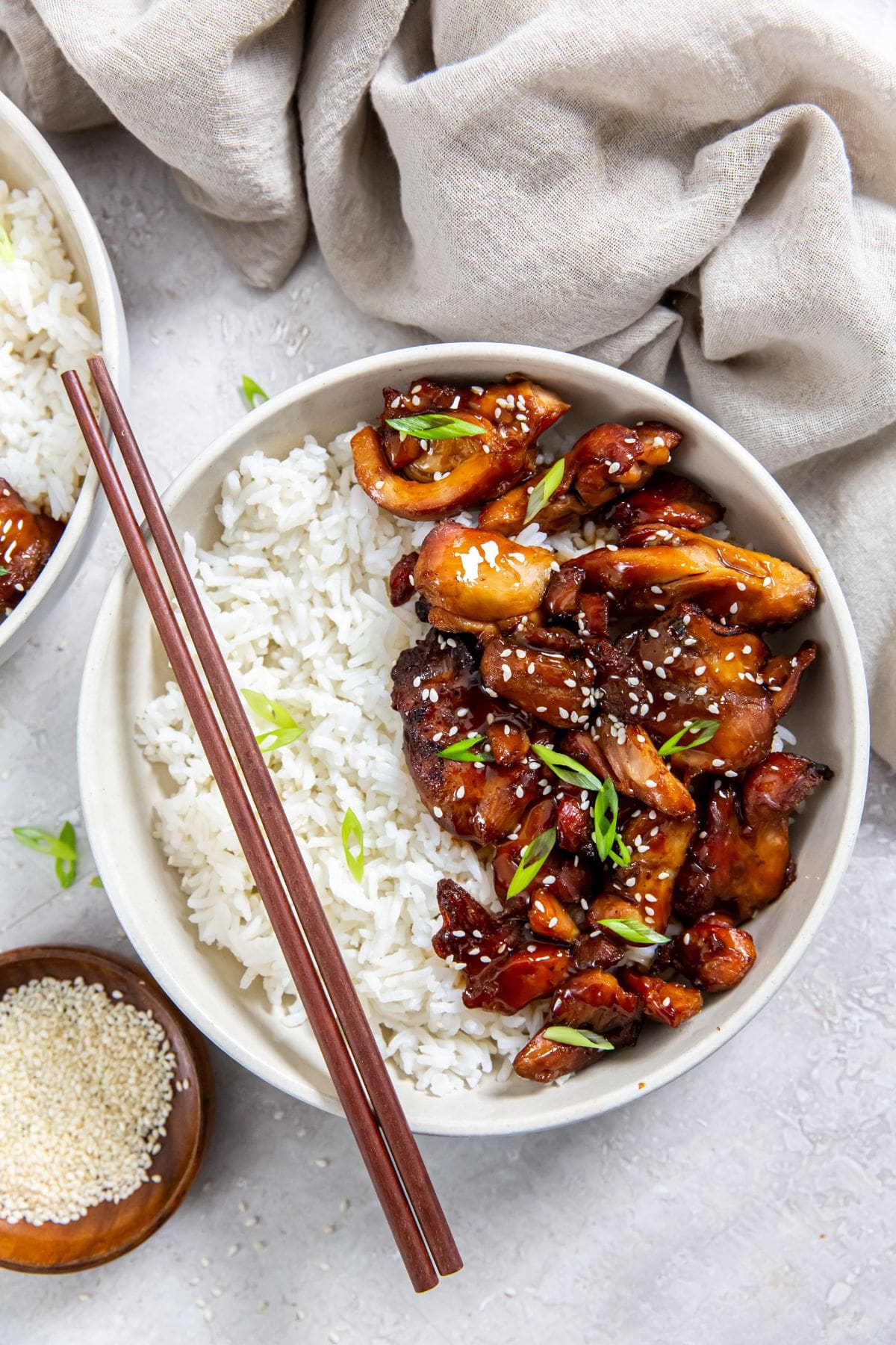 two bowls with chicken and rice inside them. gray towel in the back along with chopsticks and sesame seeds